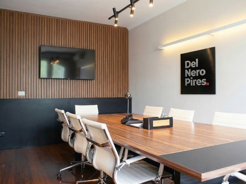 Sala de reunião elegante do escritório Del Nero Pires Advogados, mesa de madeira nobre, cadeiras brancas, painel de madeira ripada, quadro com a marca do escritório na parede, iluminação profissional, ambiente sofisticado e institucional